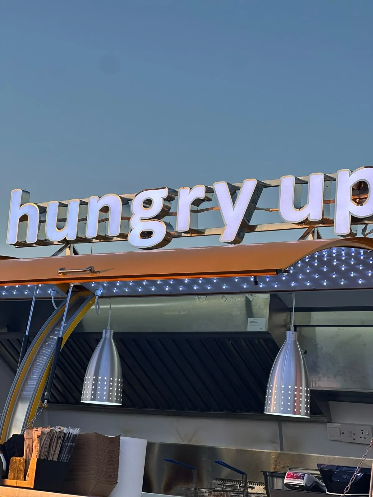 Close-up of the rooftop sign reading hungry up in rounded white letters above the open hatch, with punched steel lamps and a string of small LEDs under the awning at dusk.