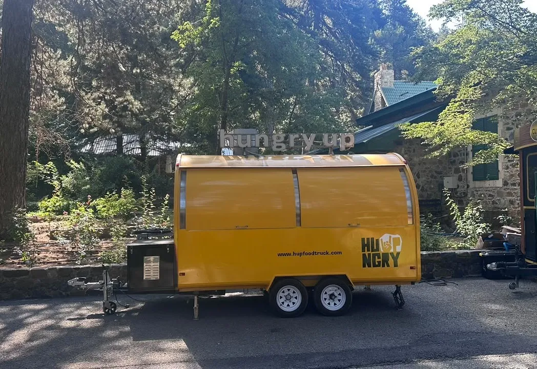 The whole Hungry Up trailer parked under pines, hitched and closed, rooftop sign above the barrel roof.