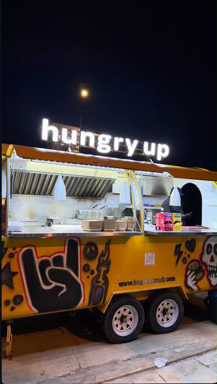 The Hungry Up trailer open and serving at night, its white rooftop sign lit, the airbrushed rock hand and skull along the yellow side.